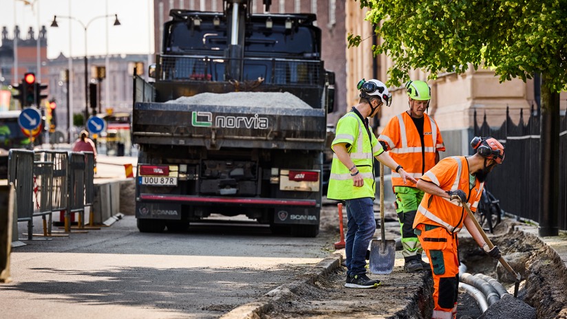 Tre män i arbetskläder arbetar i gatumiljö med en lastbil i bakgrunden