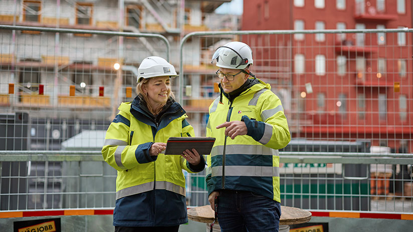 En kvinna och en man i arbetsklädsel och hjälm tittar i en ipad