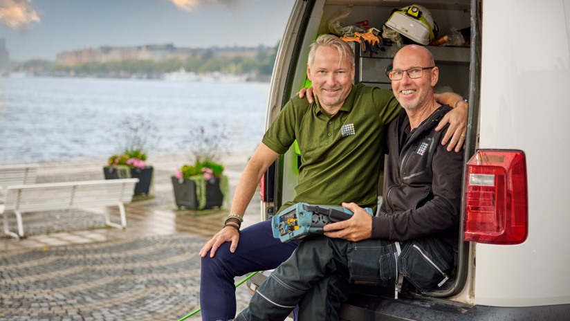 Två män sitter i bagageutrymmet på servicebil och ser glada ut med Stockholm i bakgrunden