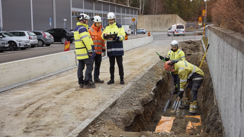 Två män gräver i ett schakt medan tre män står och på väg bredvid