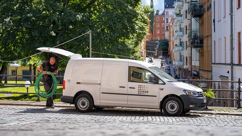 En manlig fibertekniker i arbetskläder står vid en av Stokabs vita skåpbilar en solig sommardag och plockar ut fiberkabel ur bilen. Bilen står på Södermalms kullersten med grönskande träd i bakgrunden.