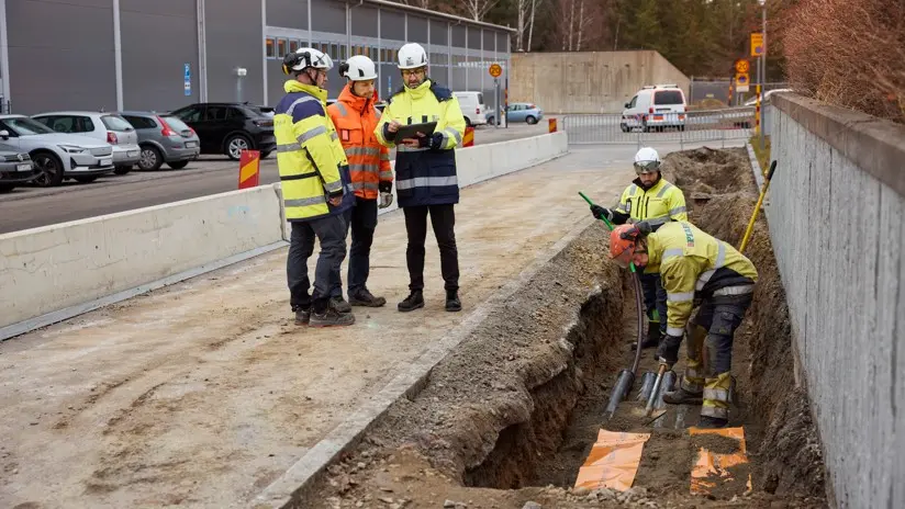 Tre män i samtal och två i schaktgrop alla bär skyddsklädsel